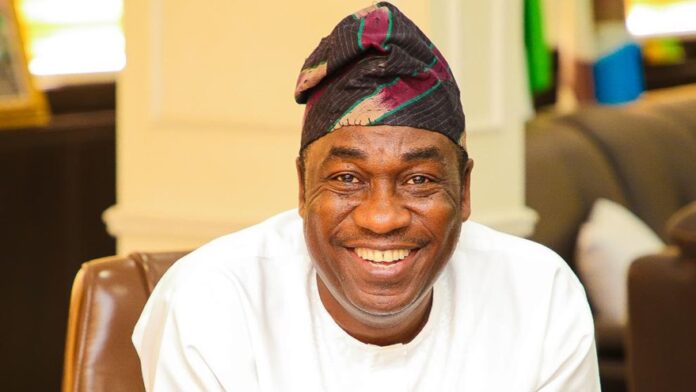 Dr.-Kadiri-Obafemi-Hamzat Smiling man in a white outfit and a patterned cap sits indoors with a warm expression.