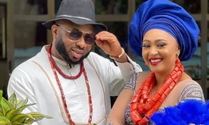 Olakunle Churchill and Rosy Meurer Couple in traditional Nigerian attire posing outdoors; man in white with hat and beads, woman in blue gele and coral beads smiling.