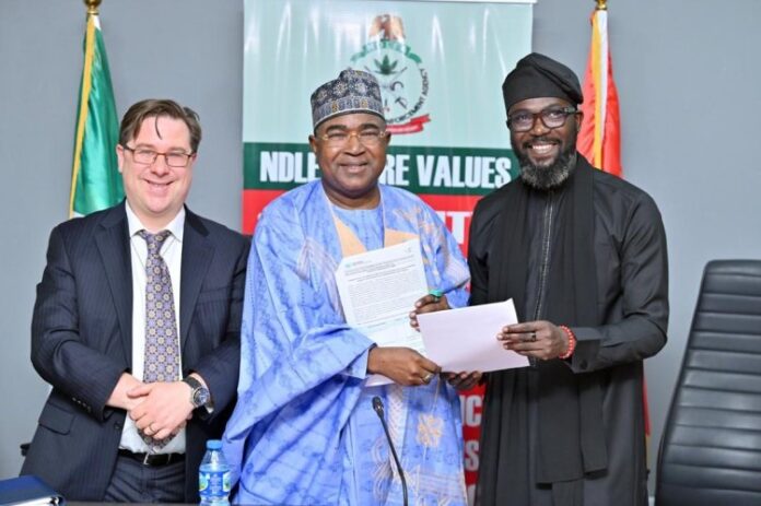 UNODC Three men at a formal ceremony: the center man in blue traditional attire holds documents while a man on his right hands him another sheet, with a suited man smiling on the left and a banner in the background.