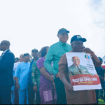 Governor Eno Unveils ARISE Agenda 2.0, Pledges More Development A diverse crowd stands outdoors at a political rally; a man in brown attire holds a campaign poster featuring Pastor Umo Eno for Governor 2027.