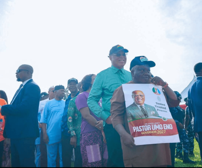 1779434170083 A diverse crowd stands outdoors at a political rally; a man in brown attire holds a campaign poster featuring Pastor Umo Eno for Governor 2027.