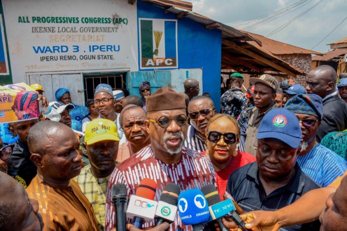 Crowd of people at a political event outside a blue building with an APC sign; multiple microphones are in the foreground for press coverage.