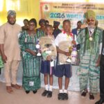 Ogun Education Commissioner Urges Students To Embrace Reading For Leadership Excellence Group of adults and two schoolchildren on a stage receiving awards, holding trophies and backpacks at a World Book Day event.