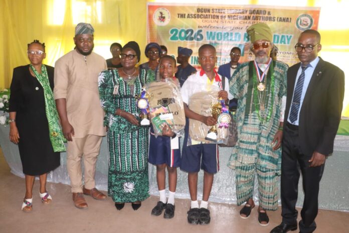 Group of adults and two schoolchildren on a stage receiving awards, holding trophies and backpacks at a World Book Day event.