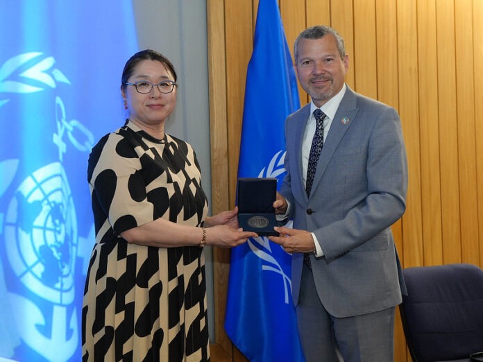 Two officials in a formal award ceremony exchanging a medal box, with a UN flag in the background.