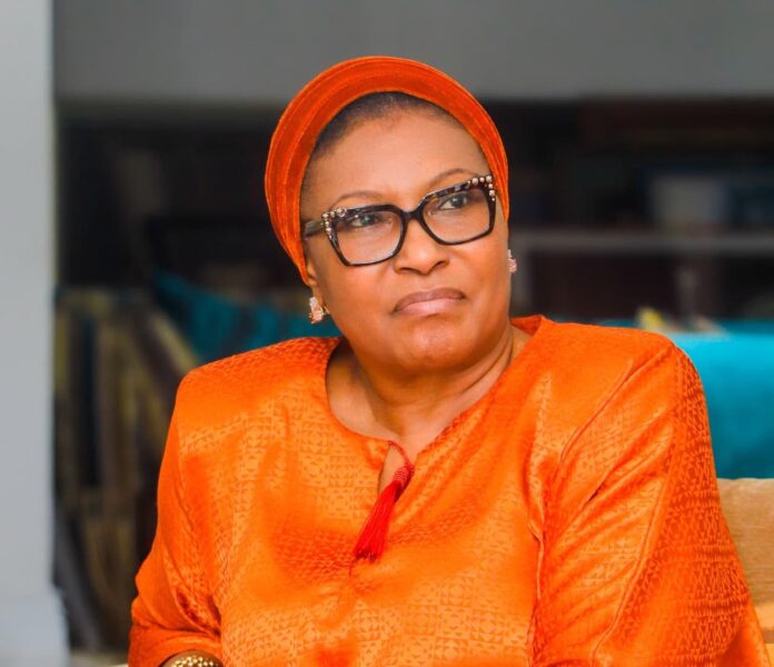 Portrait of a woman in an orange headwrap and traditional outfit, wearing glasses and looking to the side.