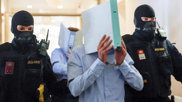7f95be30-5a8b-11f1-b4a1-c1239a456751 Two masked officers in tactical gear escort a detainee whose head is covered by a binder/box, walking through a hallway.