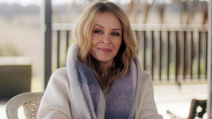 Woman with blonde hair in a light coat and gradient scarf, sitting outdoors and looking at the camera.