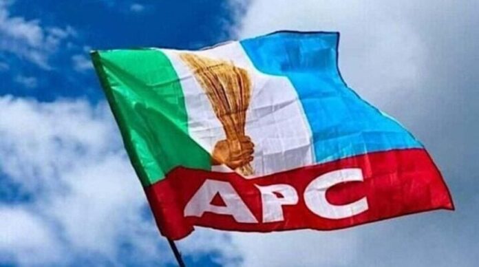 APC-flag-1024x570-1-e16841499656-860x479 Flag of the APC political party fluttering in the wind against a blue sky, featuring a torch emblem and bold 'APC' lettering on a red stripe.