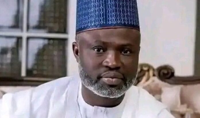 Ambassador-Abdulfatai-Yahaya-Seriki- Close-up portrait of a man with a gray beard wearing a blue patterned kufi and white traditional robes, looking at the camera indoors.