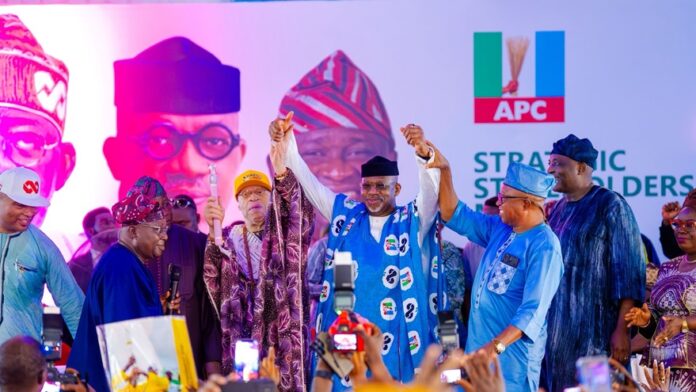 Dapo Abiodun Group of Nigerian politicians on a stage at an APC event, hands raised in celebration, audience taking photos in front of a banner with the APC logo.
