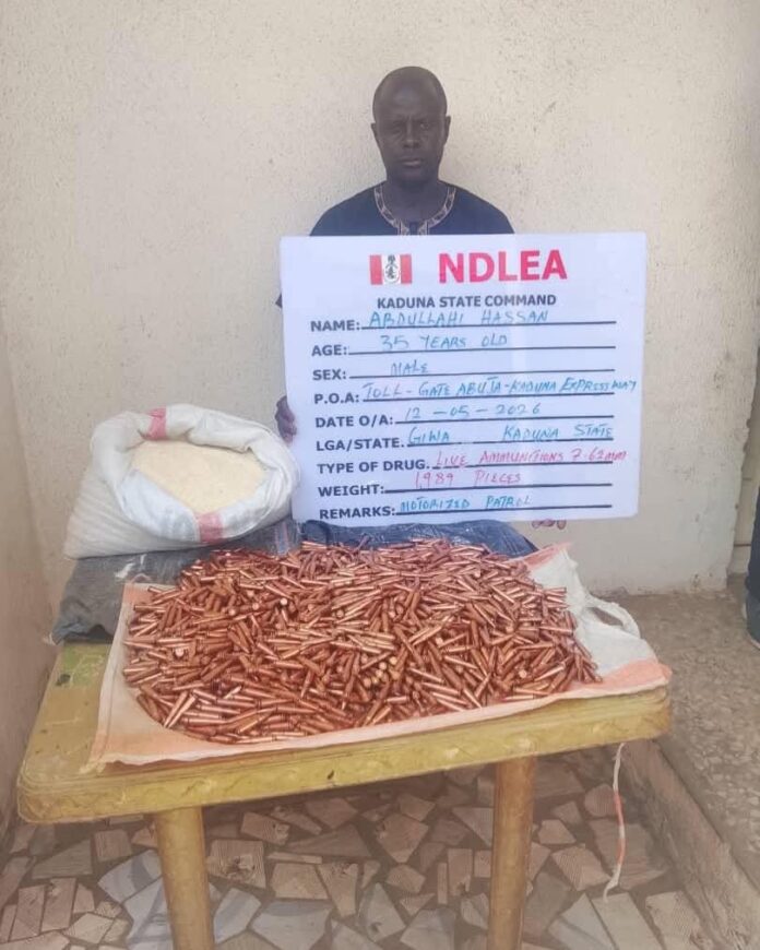 FB_IMG_1779012629208 Man sits against a wall holding a large signboard labeled 'NDLEA Kaduna State Command' with handwritten details, in front of a table piled high with many copper-colored bullets on cloth.