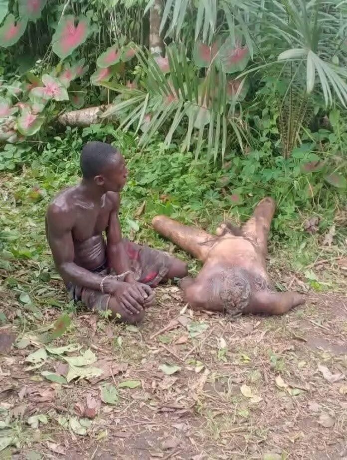 Two shirtless men in a jungle clearing; one sits on the ground as another man lies on his back among leaves and dirt, with greenery all around.
