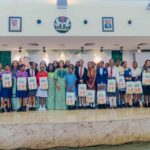No Girl Must Skip School Because Of Sanitary Pads — Oluremi Tinubu Group of students and a few adults posing on a stage indoors, each student holding a poster or brochure.