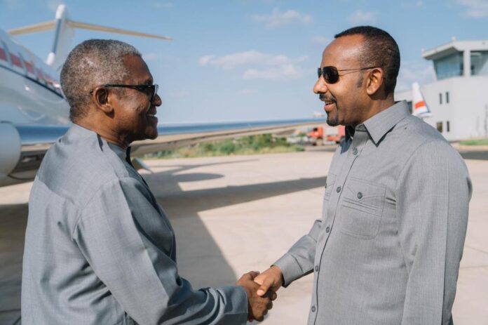 Two men in gray shirts shake hands on an airport tarmac with a jet in the background, wearing sunglasses and smiling.