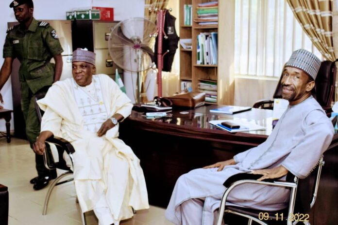 Two men in traditional Nigerian attire sit in an office across a polished desk; a guard stands in the background by a filing cabinet and fan.