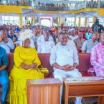 Boro Day: Governor Diri Harps On Ijaw Unity, Cooperation Front-row group at a large indoor ceremony: man in blue, woman in bright yellow dress with white headpiece, man in white, and man in pink plaid, with a crowded audience behind them.
