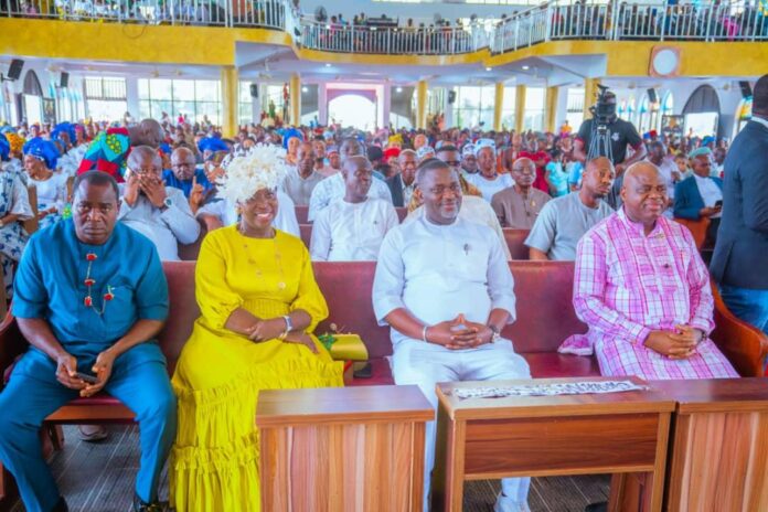 Front-row group at a large indoor ceremony: man in blue, woman in bright yellow dress with white headpiece, man in white, and man in pink plaid, with a crowded audience behind them.