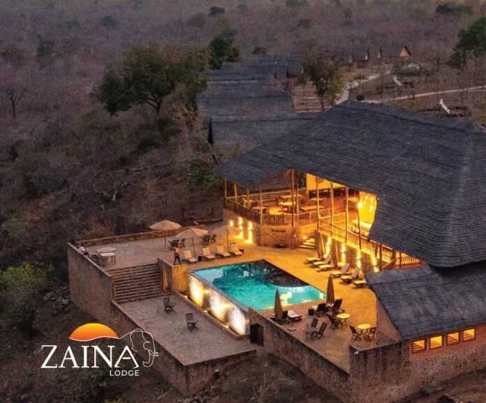 IMG-20260518-WA0017 A hillside lodge with thatched roofs, warm-lit terrace, and rectangular pool overlooking a rugged landscape; Zaina Lodge logo in the corner.