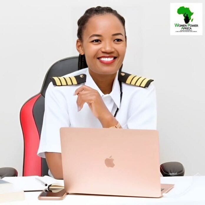 IMG-20260518-WA0066 Smiling woman in pilot uniform seated at a desk with a rose-gold laptop and papers, logo of Women Power Africa in the top-right corner.