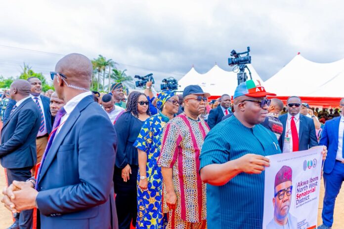Crowd at an outdoor political rally; a man in blue attire holds a campaign poster with a portrait of a candidate.