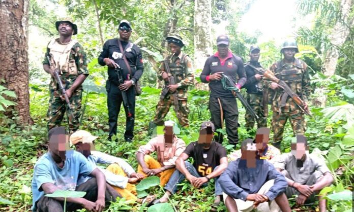 Armed men in camouflage and dark clothing stand behind a row of seated men in a forest, rifles visible.