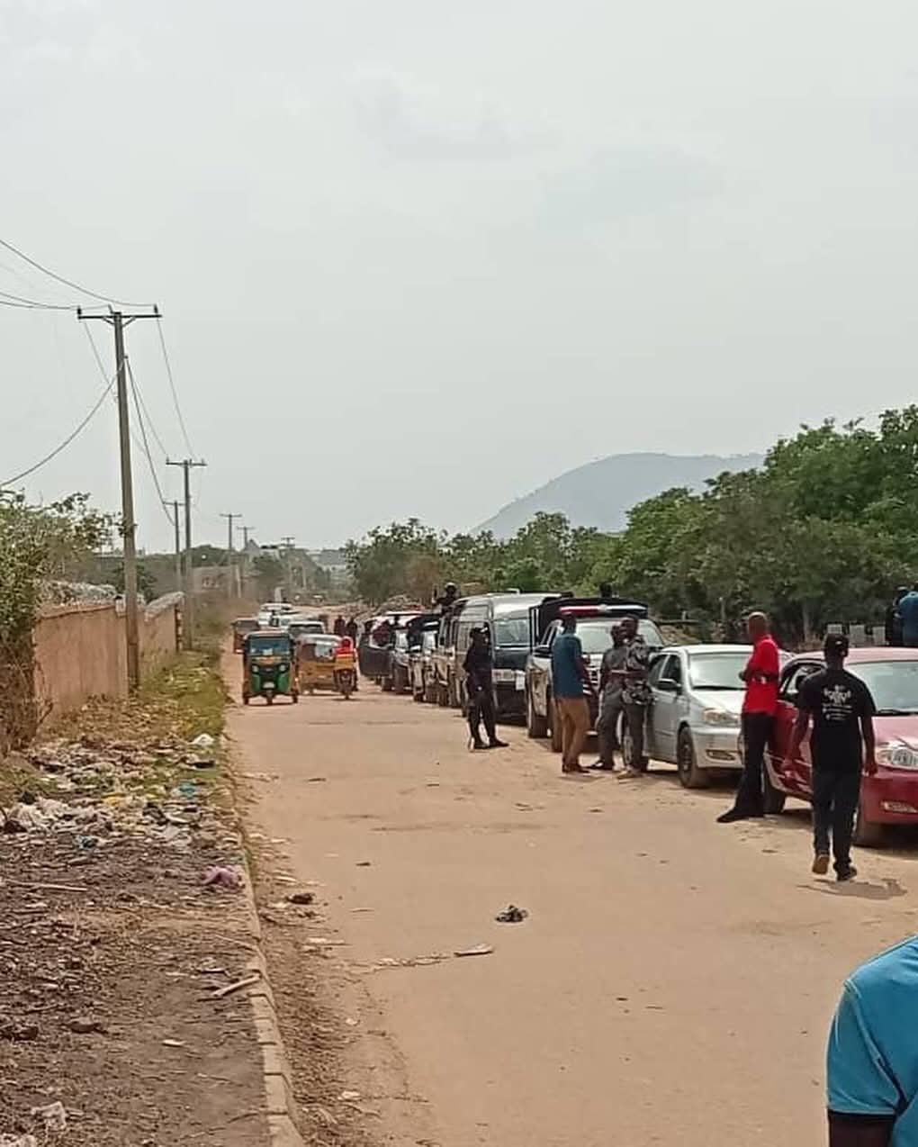IMG_5626 - THEWILL NEWS MEDIA Line of parked cars along a dusty rural street with people standing nearby and utility poles running along the road.