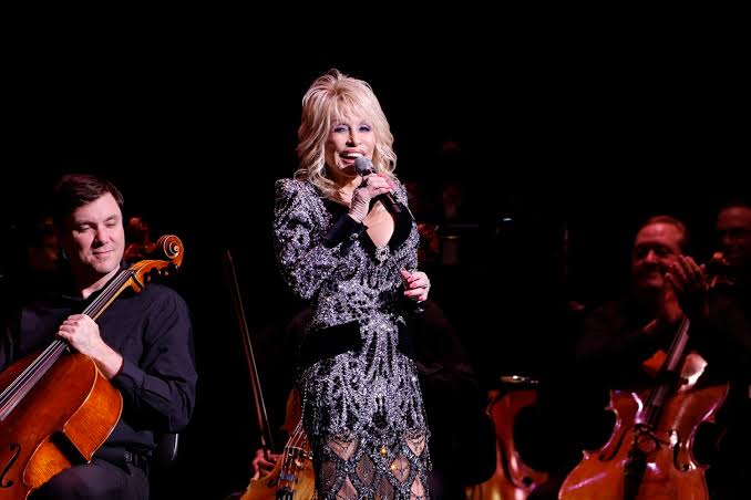 IMG_5668 Female singer in a glittering dress performing on stage with an orchestra and microphone in hand.