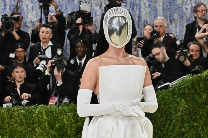 Person in a white strapless gown and long white gloves posing on a red carpet with a reflective metallic mask, surrounded by photographers.