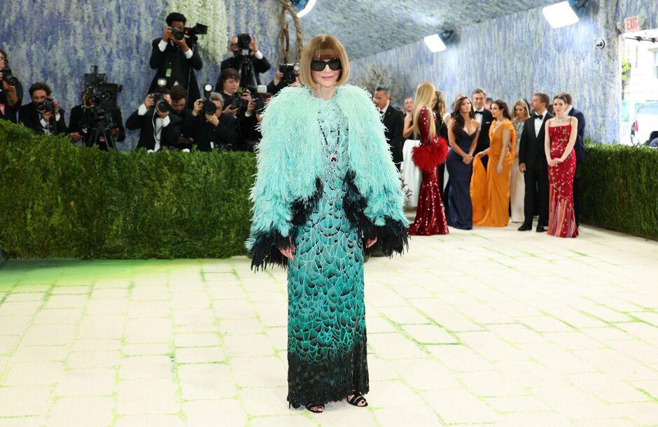 Celebrity on a red carpet wearing a turquoise feathered coat over a teal gown, posing for photographers.