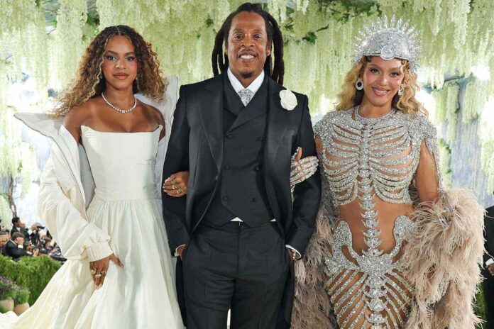 Three well-dressed individuals posing together at a formal event: a woman in a white off-the-shoulder gown on the left, a man in a black suit and vest center, and a woman in an ornate silver embellished outfit with a crown-like headpiece on the right.
