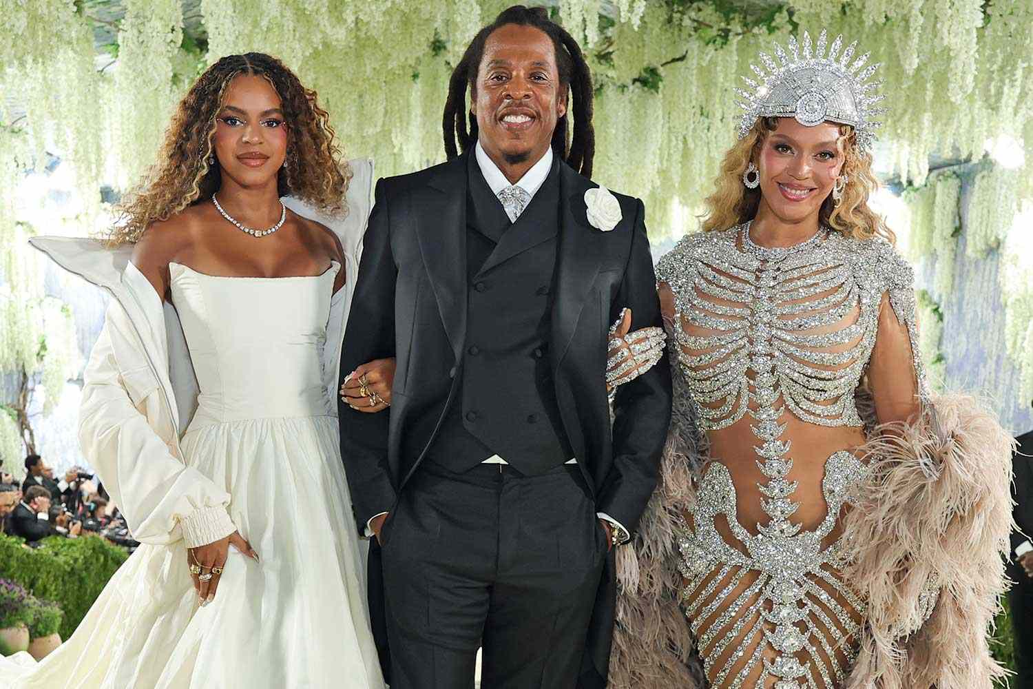 Three well-dressed individuals posing together at a formal event: a woman in a white off-the-shoulder gown on the left, a man in a black suit and vest center, and a woman in an ornate silver embellished outfit with a crown-like headpiece on the right.
