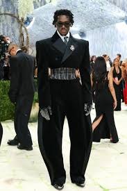 Man in an all-black, cape-style formal outfit with a wide metallic belt, gloves, and sunglasses at a red-carpet event.