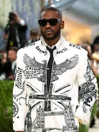 Man wearing a white graphic jacket with wings and sword designs, sunglasses, at an outdoor fashion event.