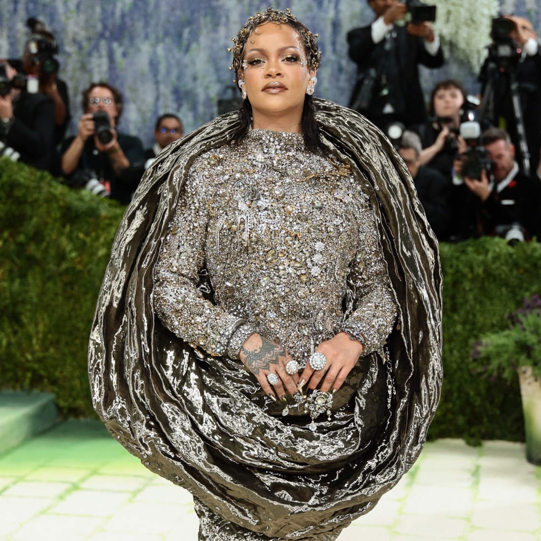 Rihanna wearing a bejeweled silver gown with a dramatic cape at a red-carpet event, photographers in the background.