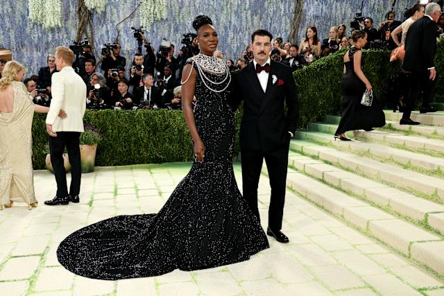 A glamorous couple poses on a light stone staircase at a red-carpet event; woman in a black sequin gown with a pearl statement necklace, man in a tuxedo.