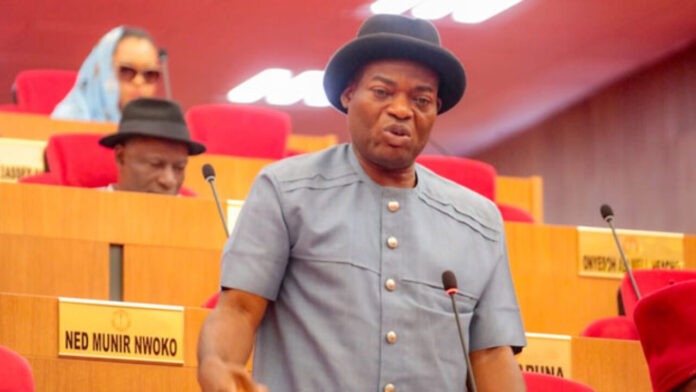 Senator Tony Nwoye Man in a gray short-sleeve suit and bowler hat speaks at a microphone in a parliamentary chamber, red seats and desks around him.