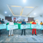 Students, Parents Hail Ime Okon As ₦16m Education Grant Reaches 320 Itu/Ibiono Beneficiaries Group of people on a stage holding oversized ceremonial checks for educational grants during a formal event inside a hall.