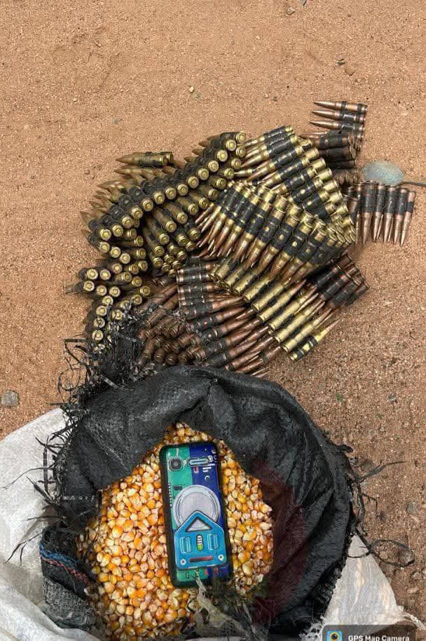 Piles of rifle ammunition on dirt beside a torn black bag spilling yellow corn and a blue handheld device.