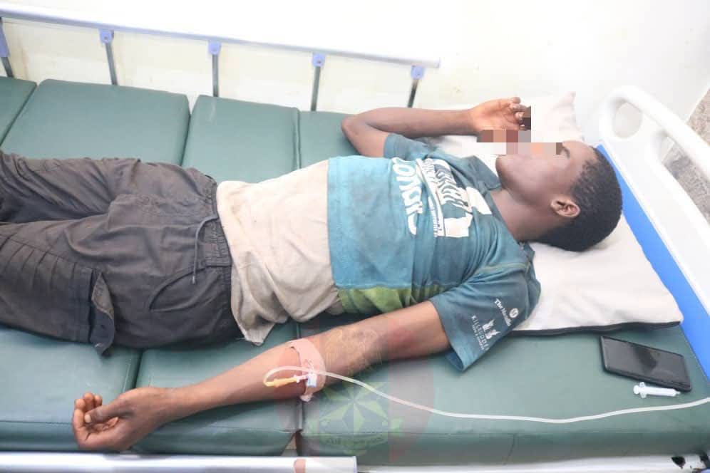 Man lying on hospital bed with IV in left arm, face pixelated, wearing a teal shirt; phone resting on bed nearby.