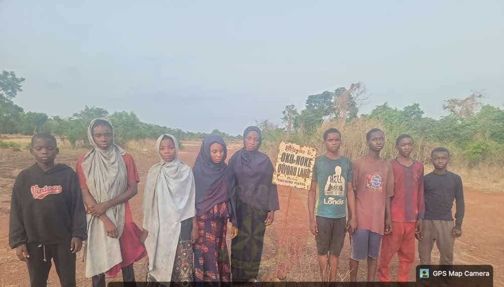 IMG_5717 - THEWILL NEWS MEDIA Group of children standing in a dusty rural field, some wearing headscarves, posing for the photo with a handwritten sign held by two kids in the center (GPS watermark bottom right).
