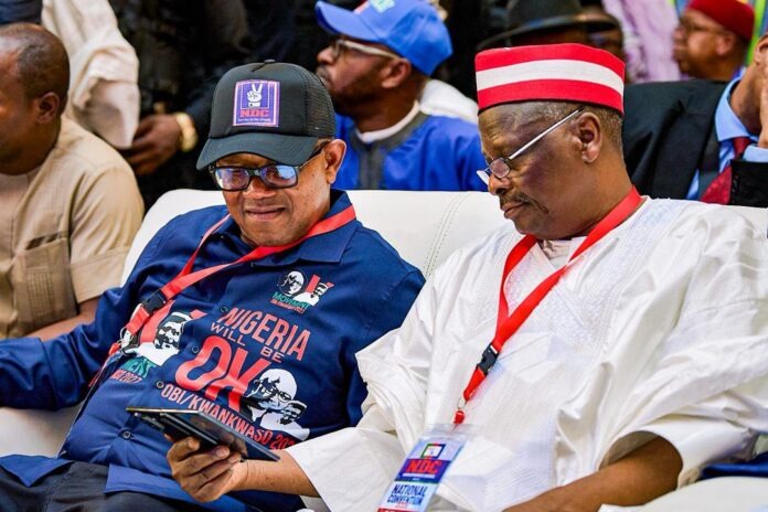 Two men sit on a white couch at a Nigerian political event; left man in blue shirt and cap looking at a tablet, right man in white traditional attire with a red cap and badge, both wearing red lanyards.