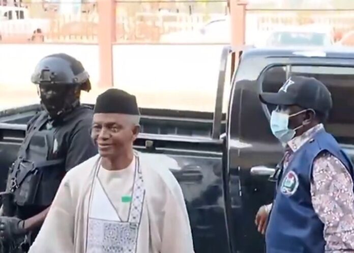 Man in a white traditional robe and black cap walks beside a dark pickup, flanked by a masked security officer in a helmet and a guard in a blue vest.