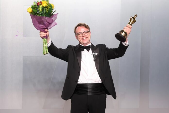 Movie Director Oscar Statuette Man in a tuxedo raises a bouquet in one hand and a golden trophy with the other, smiling at an awards ceremony.