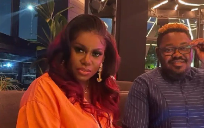 Niniola-and-her-husband Two friends sit on a couch in a dimly lit lounge, the woman wearing an orange top and long red hair, the man beside her wearing glasses and a striped shirt.