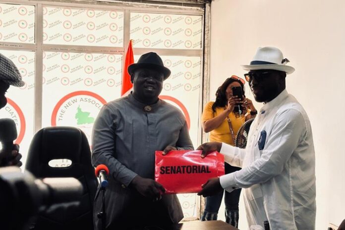 Richard Akinaka Submits PDP Nomination Form Two men in hats exchange a red pouch labeled 'Senatorial' at a formal political event, with a photographer in the background.