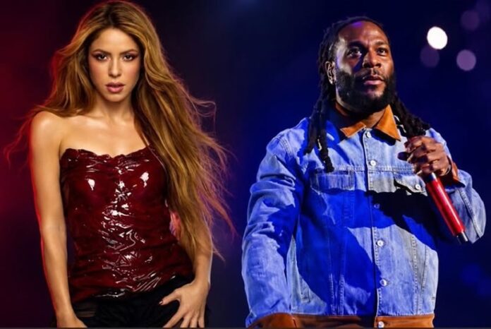 Shakira and Burna Boy Female performer in a red strapless top and black pants stands on stage next to a male rapper in a blue denim shirt, holding a red microphone, under stage lights.