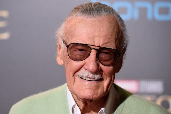 Close-up of an elderly man with gray hair and a mustache, wearing dark glasses and a pale green cardigan, smiling.