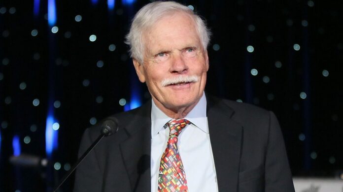 Ted Turner Senior man with white hair and mustache speaking at a podium in a dark suit and colorful tie, with stage lights in the background.