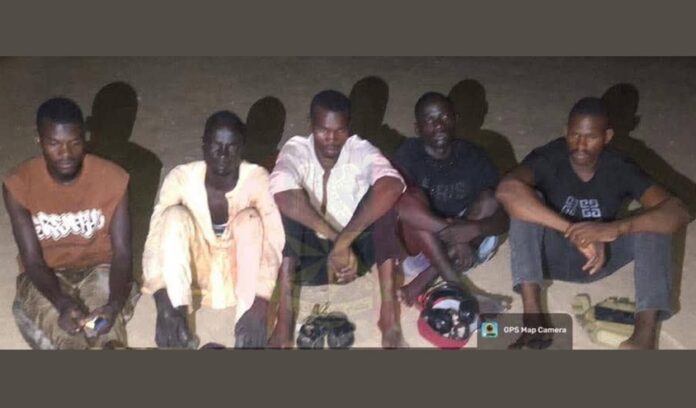 Troops Bust Terror Network Five men sit on the ground at night, posing for the photo, with large shadows cast behind them and camping gear (helmet and bags) in front.